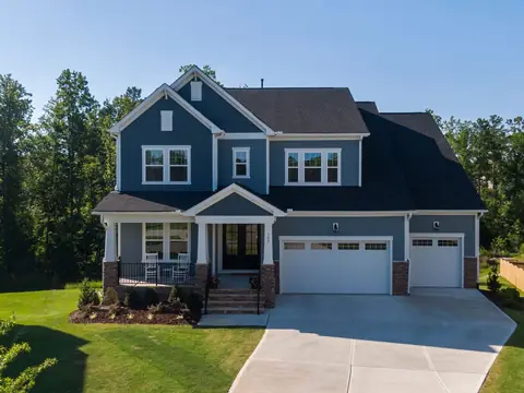 A blue two-storey house with a wide driveway and woods behind in Holly Springs, NC