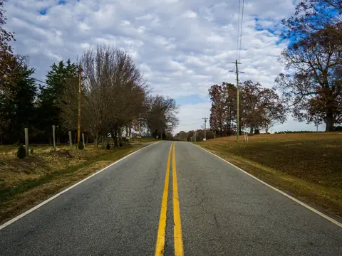 A quiet two-lane country road with yellow centre lines between autumn trees under a cloudy blue sky