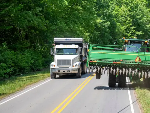 A white dump truck behind a wide green farm planter on a two-lane road lined with trees