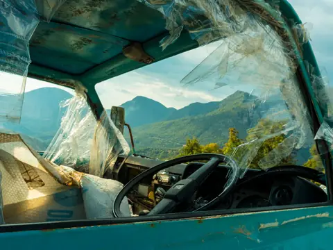 A teal car's broken side windows with a sunny mountain view through them